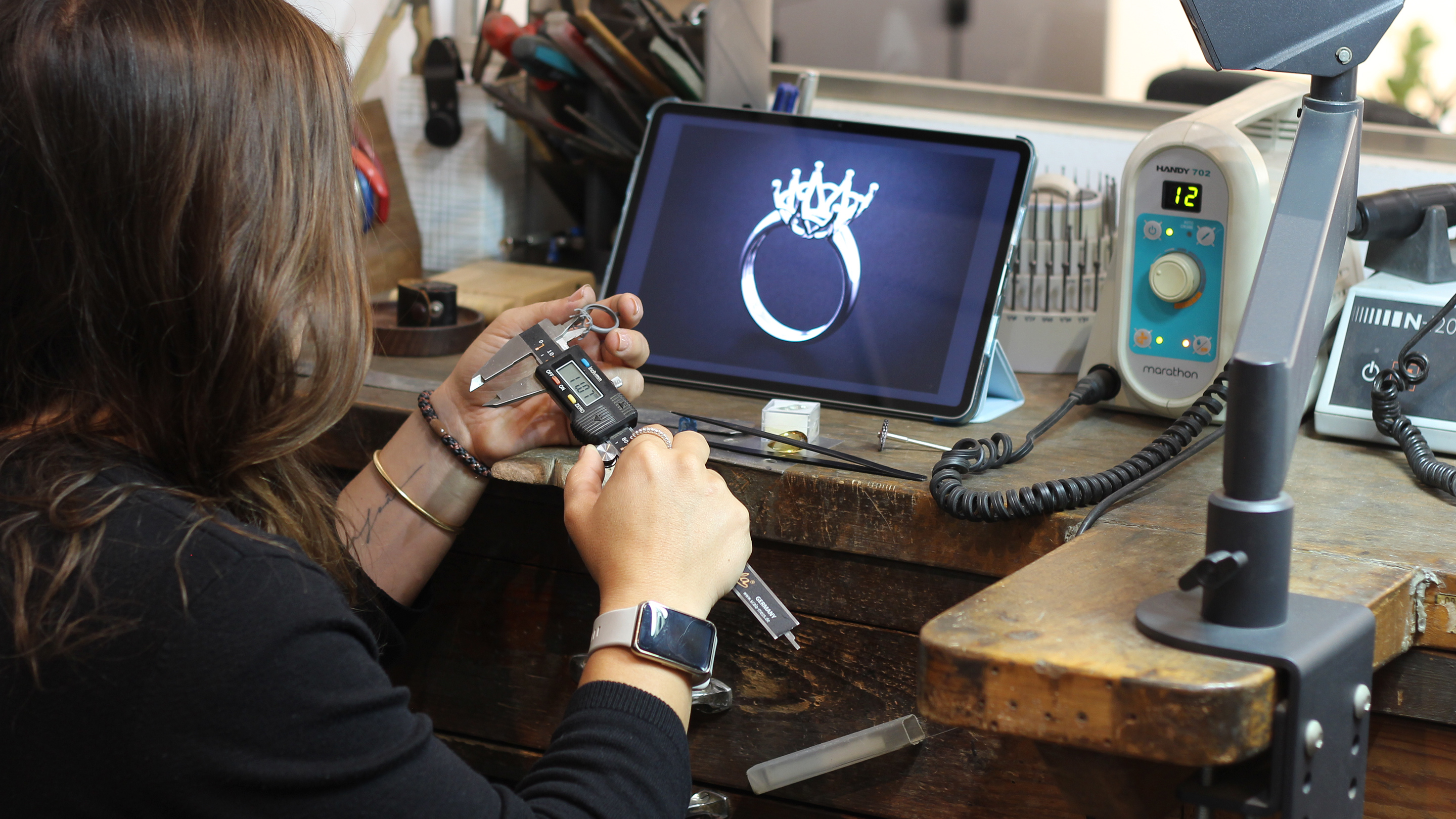 Diana en un banco de madera comprobando un prototipo con un calibre, mientras una tablet muestra el render CAD de la misma pieza